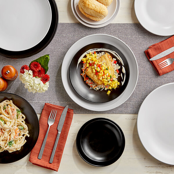 A black Acopa stoneware coupe plate on a table with food and a white plate.