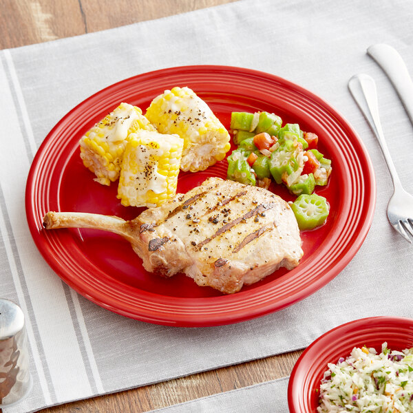 An Acopa Capri red stoneware plate with a pork chop and corn on the cob on a table.