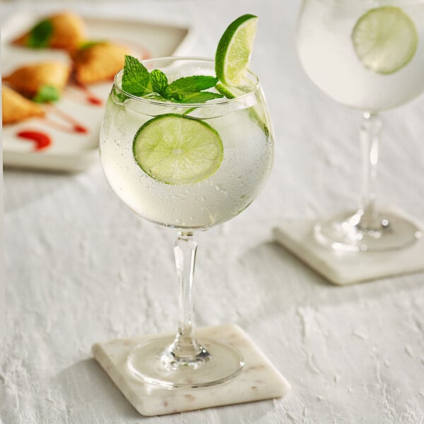 Two Acopa Empire gin and tonic cocktail glasses with white liquid and lime slices on a table.