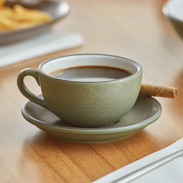 A moss green matte stoneware cup filled with coffee, placed on a matching saucer with a rolled wafer stick.