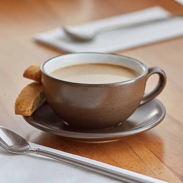 A close-up of an Acopa Embers hickory brown matte stoneware cup of coffee on a saucer with a cookie.