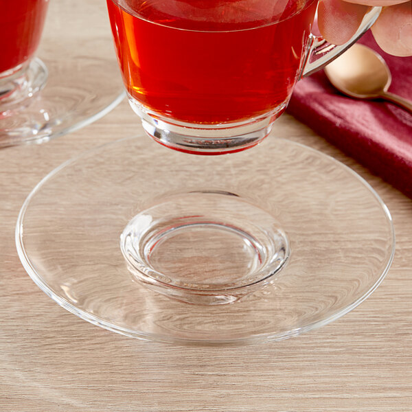 A hand holding an Acopa coffee saucer with a glass cup of red liquid.