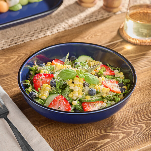 A blue stoneware coupe low bowl filled with a fresh salad containing strawberries, blueberries, corn, cucumber, and greens.