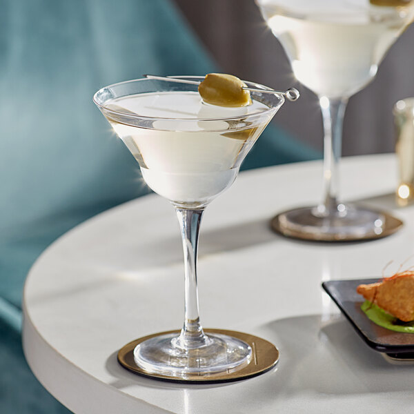 Two Acopa martini glasses with olives on top of them on a table in a cocktail bar.