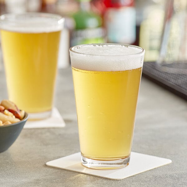 Two Acopa highball glasses of beer on a counter with a bowl of nuts.