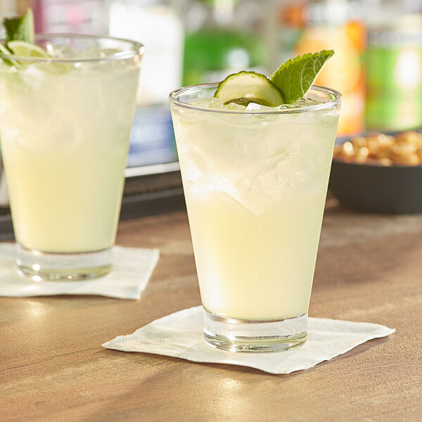 Two Acopa Fusion beverage glasses filled with lemonade garnished with mint and cucumber on a table.