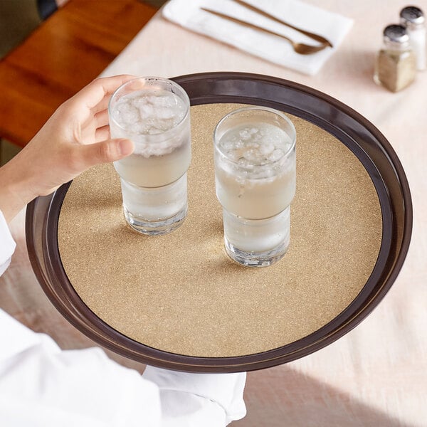 A hand holding a Carlisle brown cork-lined serving tray with two glasses of water.