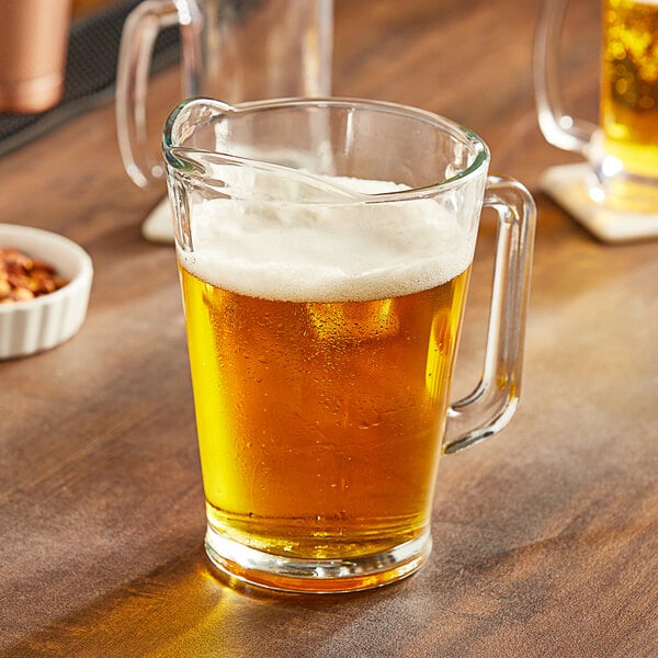 An Acopa glass beer pitcher on a table in a bar full of beer.