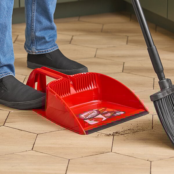 A person using a Libman red dust pan with a broom to sweep a wood floor.