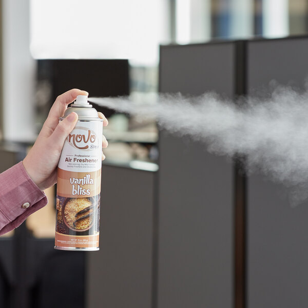 A person spraying Noble Chemical Novo Vanilla aerosol air freshener in a corporate office cafeteria.