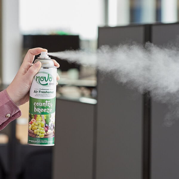 A hand holding a green and white aerosol can of Noble Chemical Country Breeze air freshener spraying it in a room.