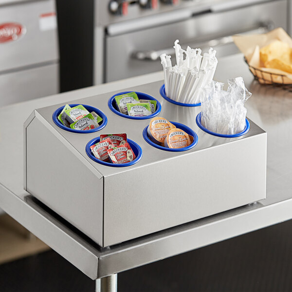 A stainless steel flatware organizer with blue perforated plastic cylinders on a counter with condiments and straws.