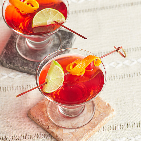 Two glasses of red liquid with lime slices and orange peels with a red bamboo knot skewer in one glass.