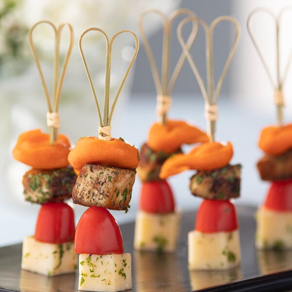 Bamboo skewers with heart-shaped ends holding cheese, tomatoes, and orange food items.