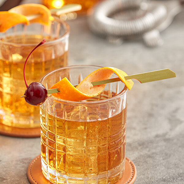 Two glasses of alcohol with a Bamboo Paddle Skewer holding an orange peel and a cherry.