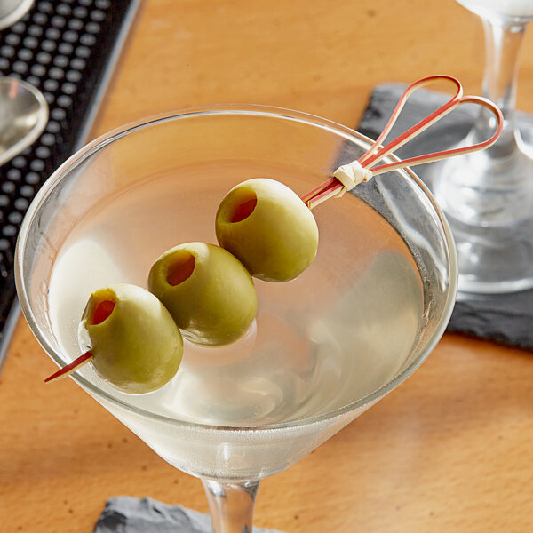 A martini glass with a red bamboo heart skewer holding green olives.