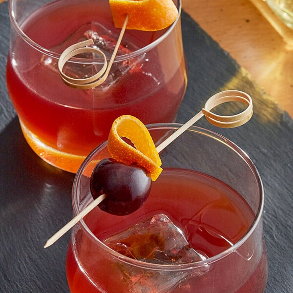 Two glasses of red liquid with orange slices and a cherry on a Bamboo Loop Food Pick on a table in a cocktail bar.