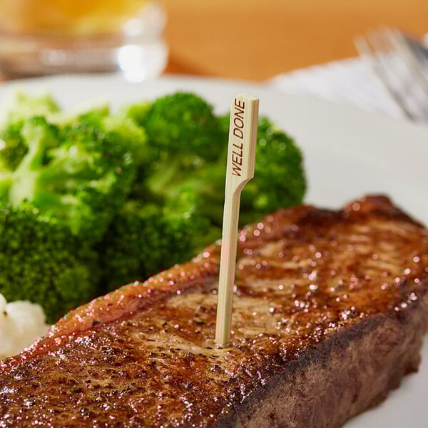 A steak with a Bamboo by EcoChoice "Well" Meat Marker in it sitting on a plate of broccoli.