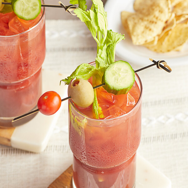 A glass of red liquid with a cucumber and green olive garnish with a black bamboo skewer.