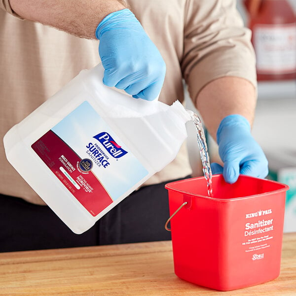 A person in blue gloves pouring Purell fragrance-free surface sanitizer into a bucket.