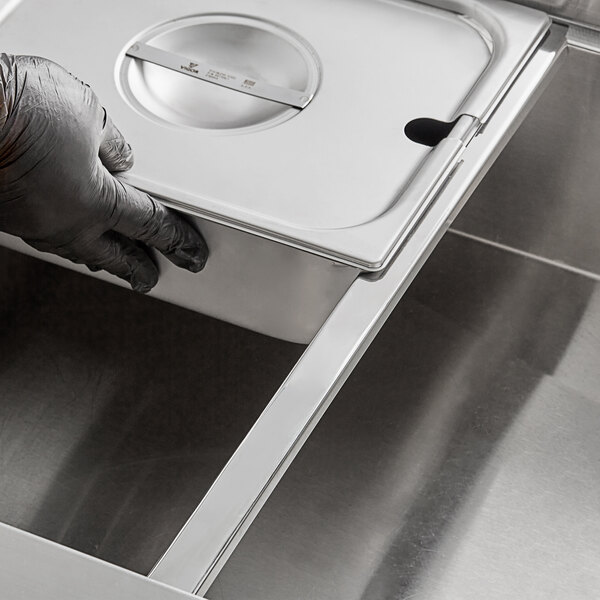 A person wearing gloves using a Vigor steam table adapter bar to hold a metal container.