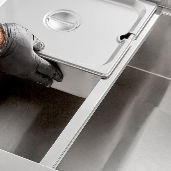 A person in black gloves using a Choice stainless steel hotel pan adapter bar to hold a metal container over a sink.