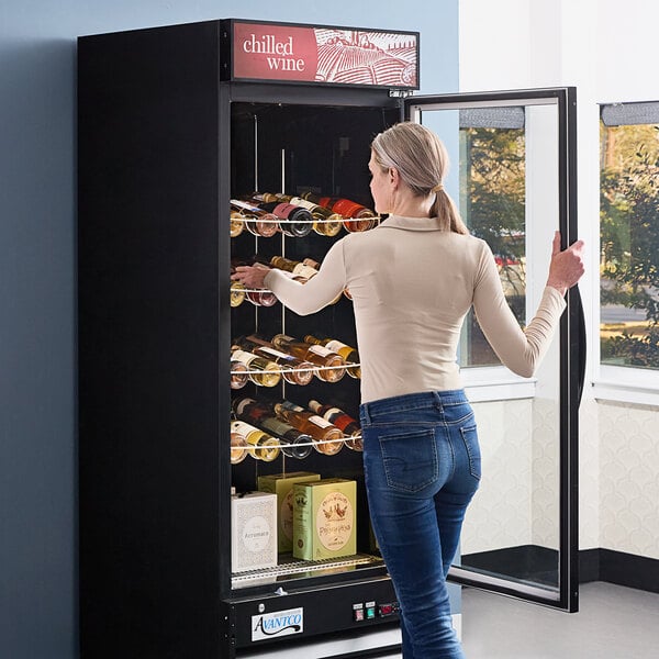 A woman looking at an Avantco black single door wine merchandiser filled with wine bottles.