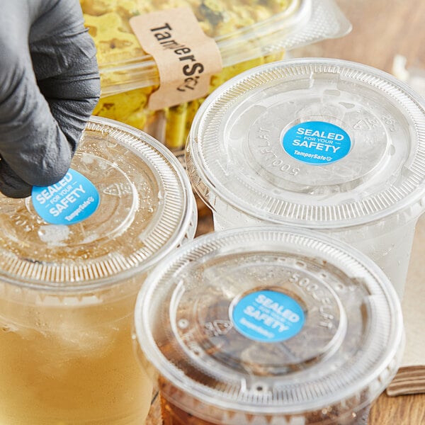 A gloved hand applies a blue TamperSafe label to a plastic cup.