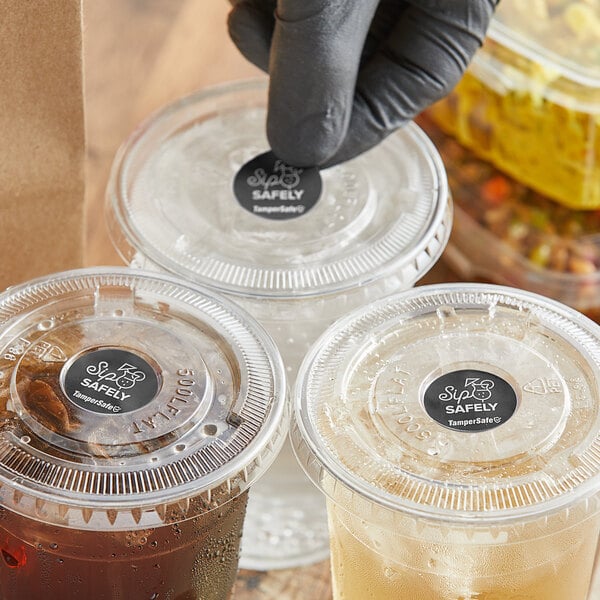 A hand in a black glove placing a TamperSafe paper label on a plastic cup with a lid containing a drink.