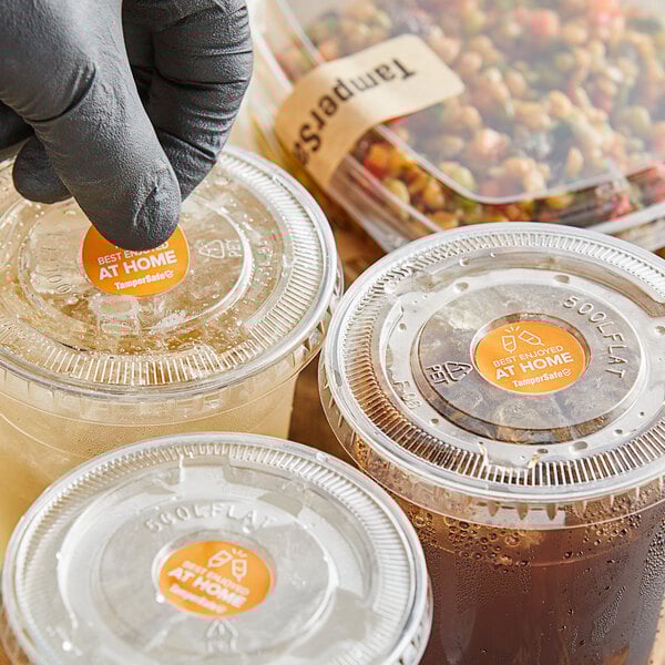 A gloved hand putting a TamperSafe orange label on a plastic cup of brown liquid.