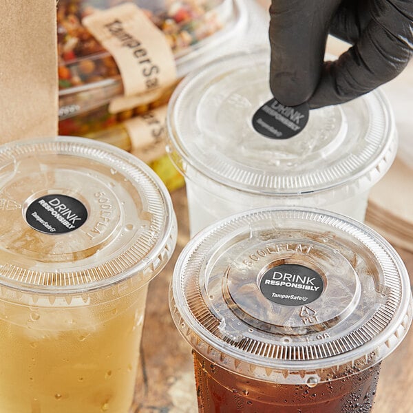 A hand in a black glove applying a TamperSafe round drink label to a plastic cup with a lid and a drink inside.