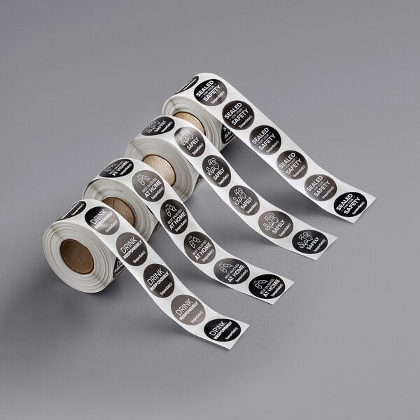 A roll of TamperSafe black and white drink labels with white circles and words on a gray surface.