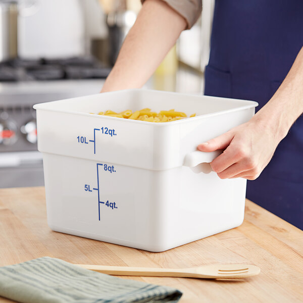 A white square 12-quart polypropylene food storage container with blue measurement markings, being held on a kitchen counter.