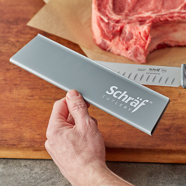 A hand holding a Schraf knife with a gray polypropylene blade guard over a piece of meat on a counter.