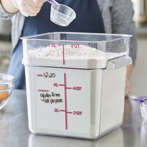 A woman using a measuring cup to pour flour into a Carlisle square clear plastic food storage container.