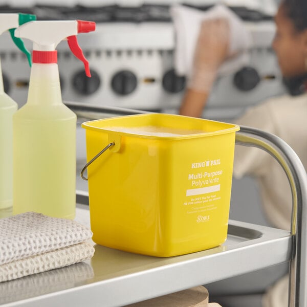 A yellow Noble King-Pail 3 quart cleaning pail on a cleaning cart next to spray bottles and cleaning cloths.