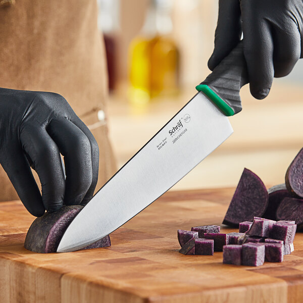 A person wearing black gloves uses a Schraf chef knife with a green handle to cut a purple potato on a counter.