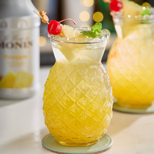 A glass pitcher filled with yellow Monin Pineapple Fruit Puree on a table with two glasses of yellow drinks.
