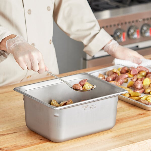 A person in a chef's uniform using a spoon to serve food from a Vollrath stainless steel hotel pan.