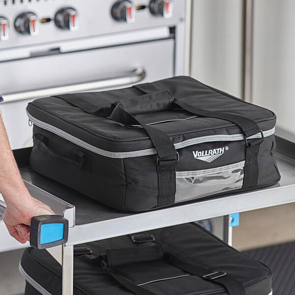 A man holding a black Vollrath insulated snack delivery bag with a strap and two buckles.