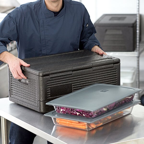 A man in a chef's uniform holding a black CaterGator food pan carrier with a lid.