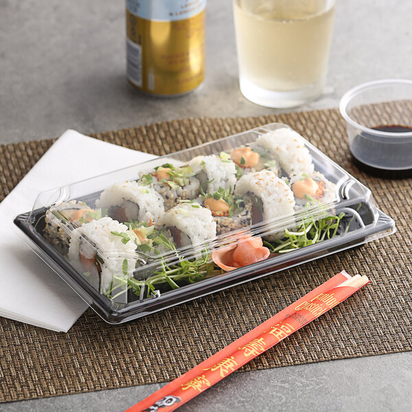 A clear plastic extra large food container with a lid, holding sushi rolls, displayed on a table with chopsticks and a drink.
