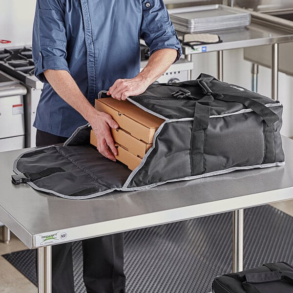 A man in a chef's uniform holding a Vollrath insulated pizza delivery bag with pizza boxes inside.