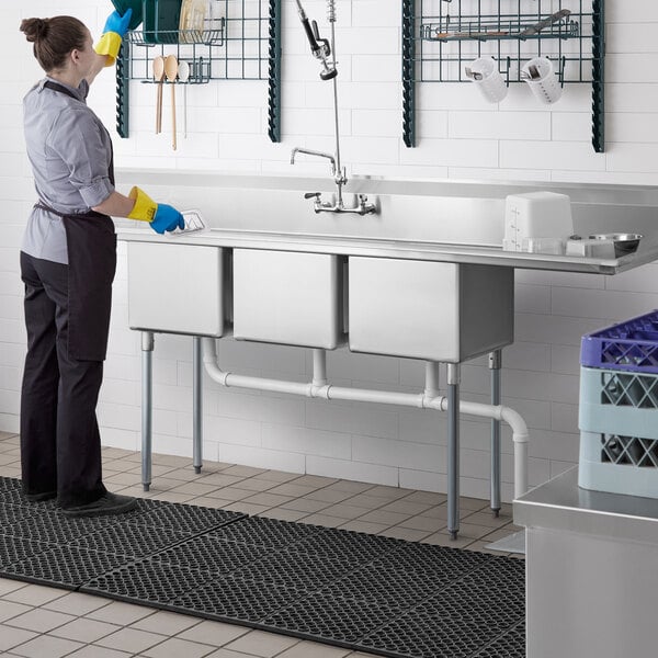 A woman in a professional kitchen wearing an apron and gloves washing a Regency 3 compartment sink.