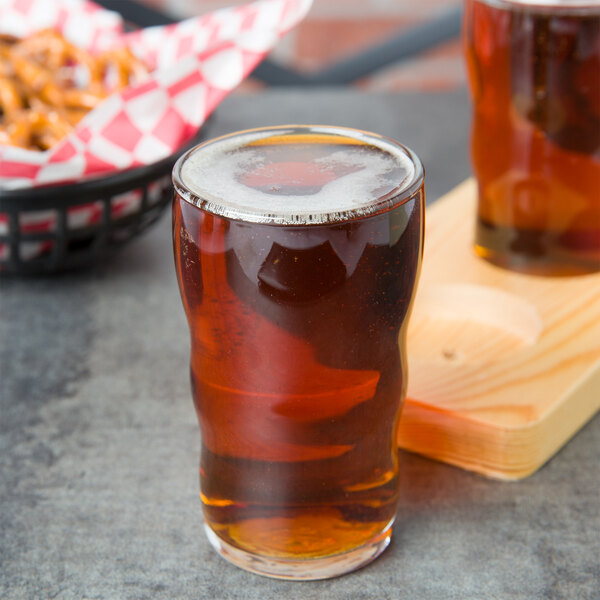 A Libbey juice glass filled with beer next to a bowl of pretzels on a table.