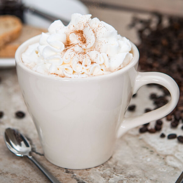 A Tuxton Europa eggshell cappuccino mug filled with coffee and whipped cream with a spoon next to it.