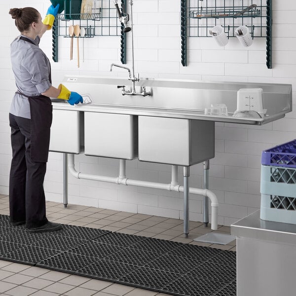 A woman in a blue shirt and yellow gloves cleaning a Regency 3 compartment sink on a counter in a professional kitchen.