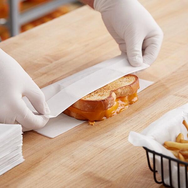 A hand wearing white gloves uses a knife to cut a grilled cheese sandwich on dry wax paper.