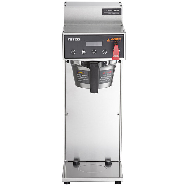 A silver Fetco airpot coffee brewer with a black handle and metal brew basket on a stainless steel stand.