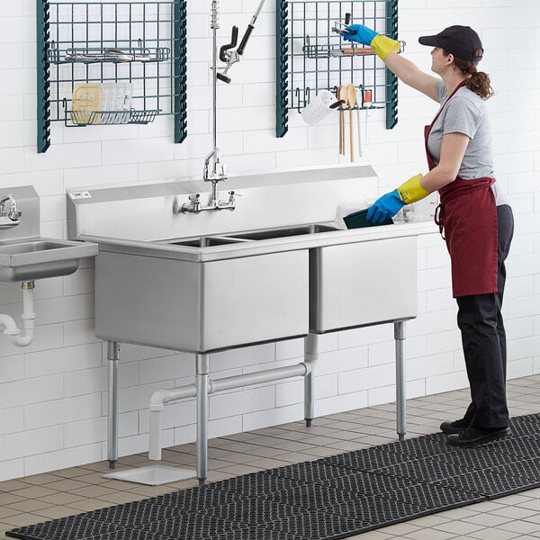 A woman in a black apron cleaning a Regency stainless steel two compartment sink in a professional kitchen.